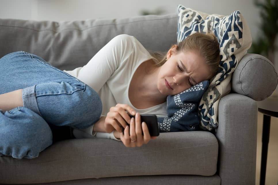 Kobieta leży na łóżku z telefonem w ręku ze smutną twarzą, zastanawiając się jak sobie poradzić z trudnymi relacjami w swoim życiu.